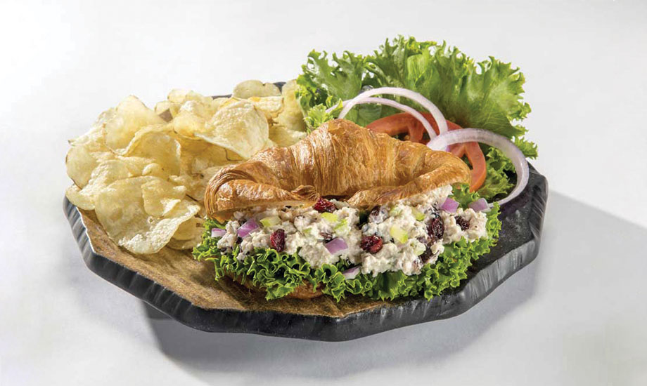 Image of chicken salad sandwich on a croissant served with potato chips, lettuce, onion and tomato on the side
