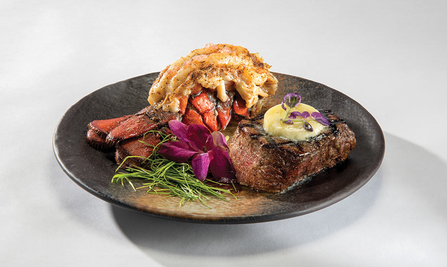 Image shows surf and turf dish with a filet and a lobster tail and magenta flower garnish