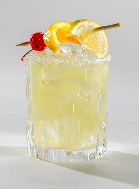 Image shows a yellow colored cocktail with cherry, orange and lime garnish