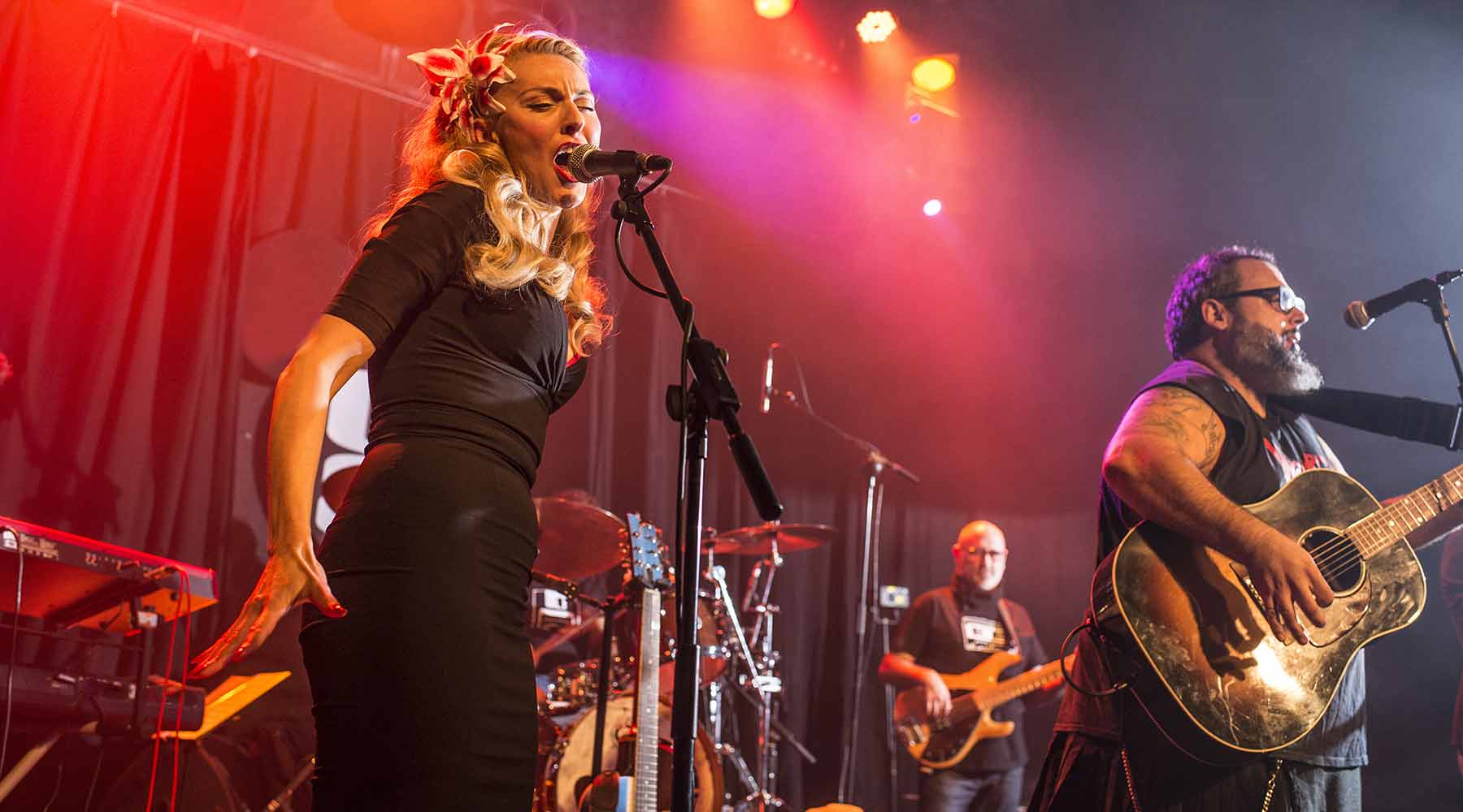 Image shows blond woman signing into a microphone on red-lit stage with other band members playing music around her