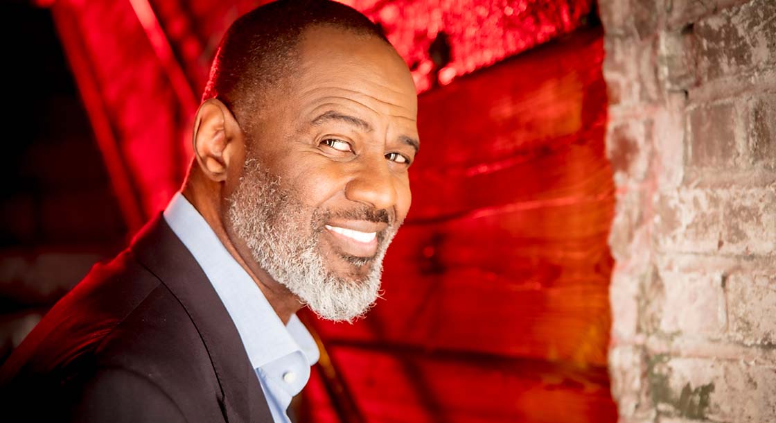 Image shows singer Brian McKnight facing right and looking at the camera, with a red background behind him